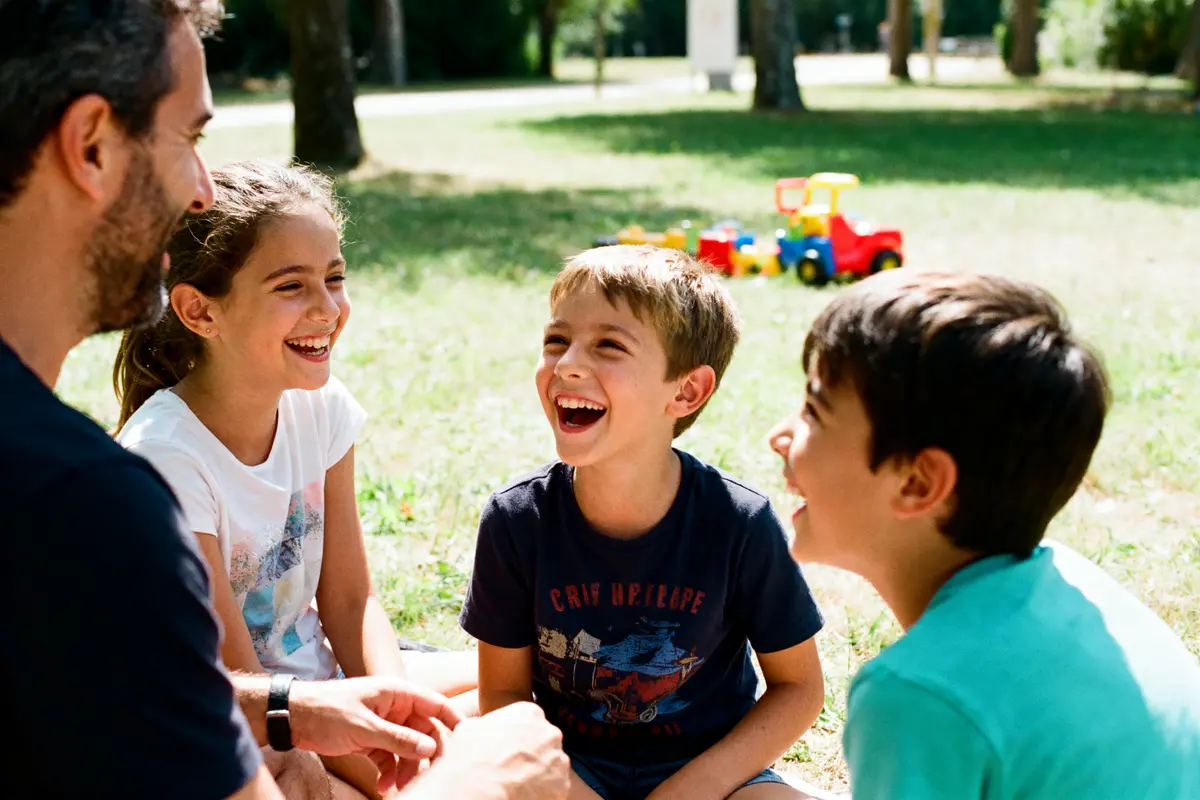pexels-michal-pech-213601-757318 (1) Un adulte et trois enfants riant ensemble, assis dans un parc ensoleillé, jouets colorés en arrière-plan.