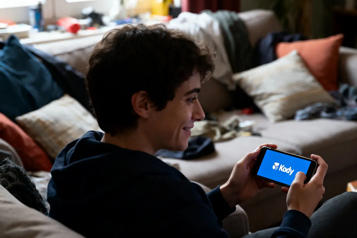 pexels-michal-pech-213601-757318 (1) Jeune homme souriant tenant téléphone affichant logo "Kody", assis dans un salon avec des coussins colorés.