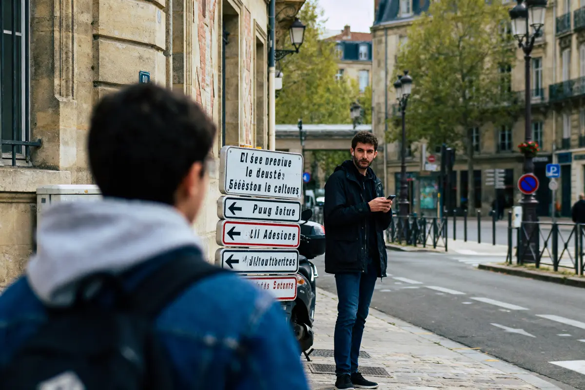 Deux hommes dans une rue urbaine, panneaux de direction illisibles, bâtiments historiques, arbres en arrière-plan.