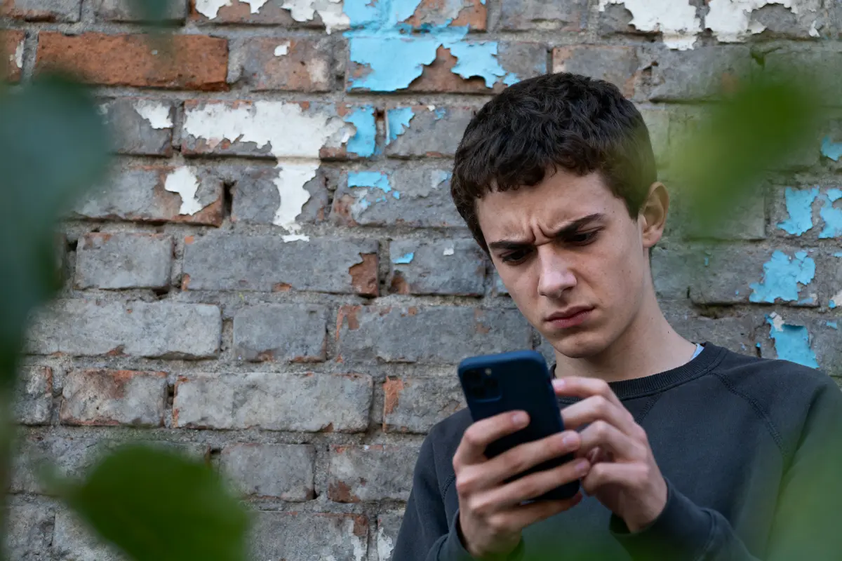 pexels-michal-pech-213601-757318 (1) Un jeune homme sérieux regarde son téléphone devant un mur de briques usé, avec des éclats de peinture bleue.