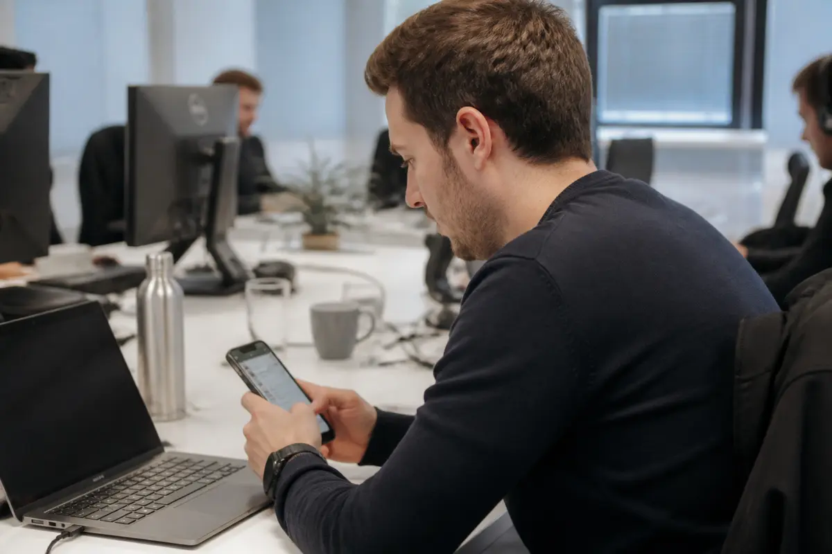 Un homme utilise un smartphone en milieu de travail, ordinateur portable et bureau en arrière-plan.